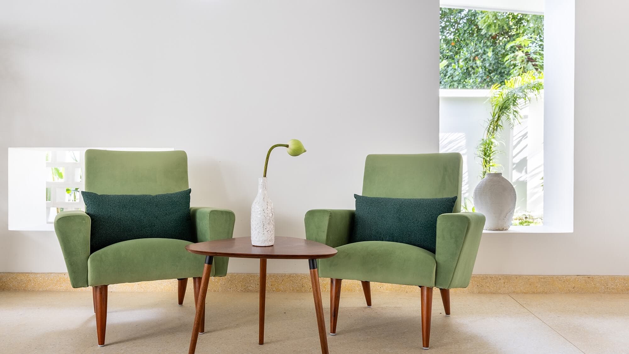 Two green armchairs with dark cushions face a small wooden table holding a white vase with a single flower, creating an inviting corner that reflects the elegance of Siem Reap Luxury Accommodation against large windows and a white wall.