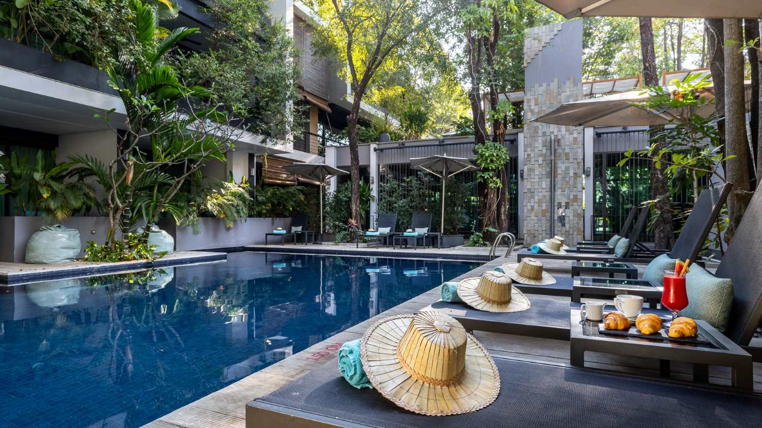 Anjali by Syphon luxurious poolside area with sun loungers, straw hats, and a breakfast setup in Siem Reap, surrounded by lush greenery and modern architecture.