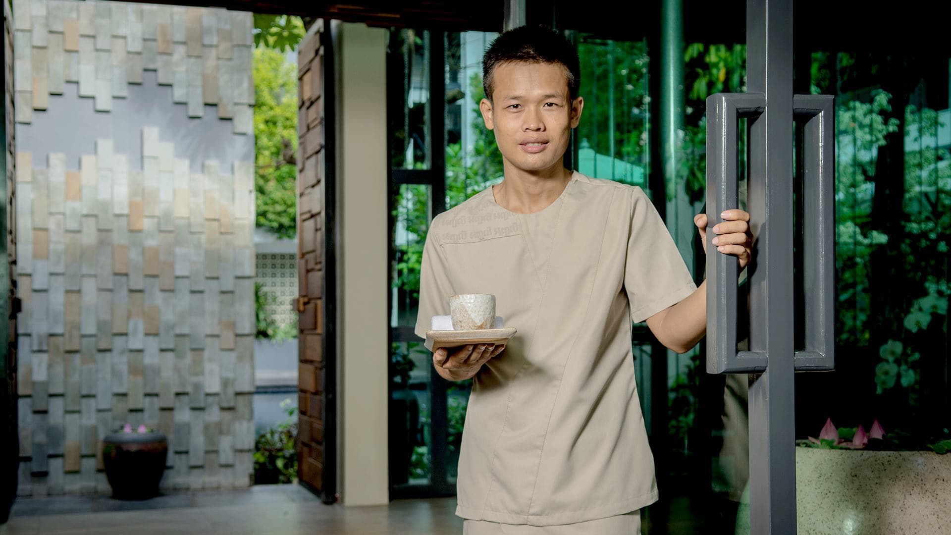 A man in a beige uniform holds a tray with a cup and saucer, standing by an open door in a serene, garden-like setting in Siem Reap.
