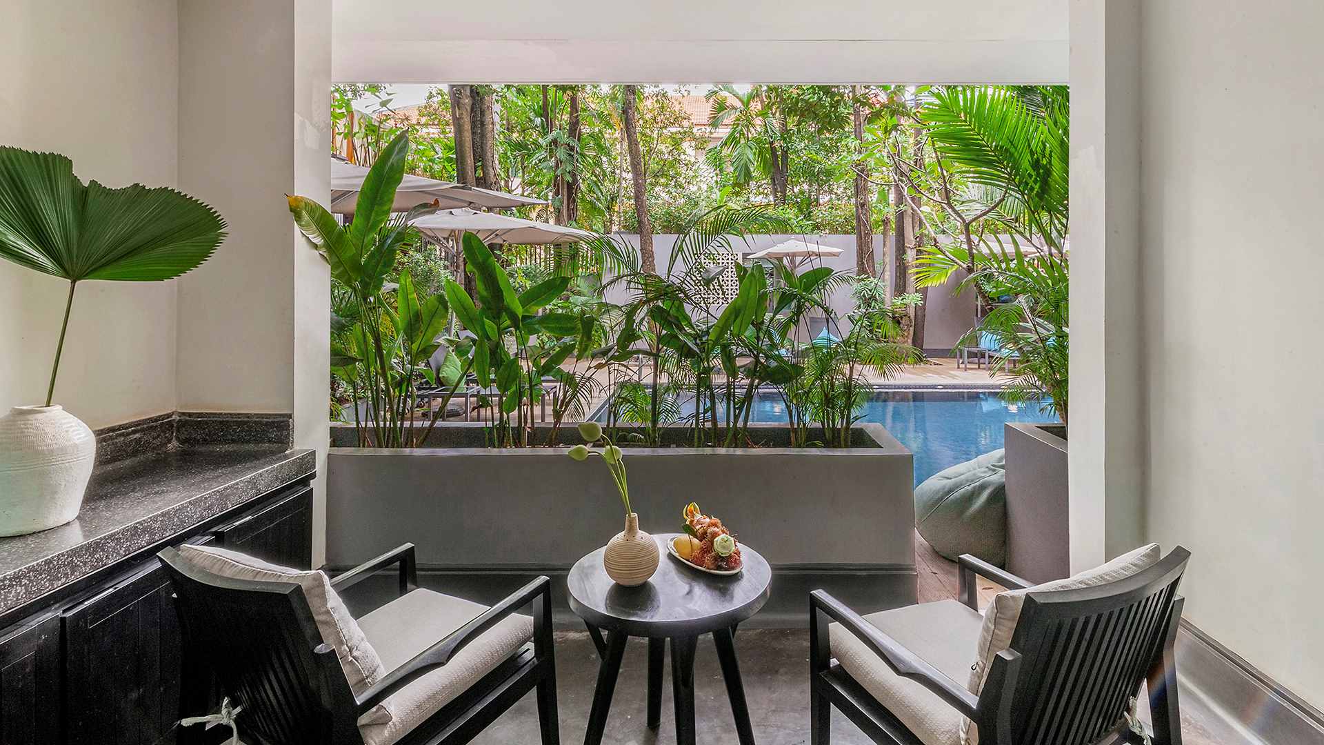 View from an open window showing a pair of chairs and a table overlooking a serene hotel pool in Anjali by Syphon, Siem Reap surrounded by lush greenery.