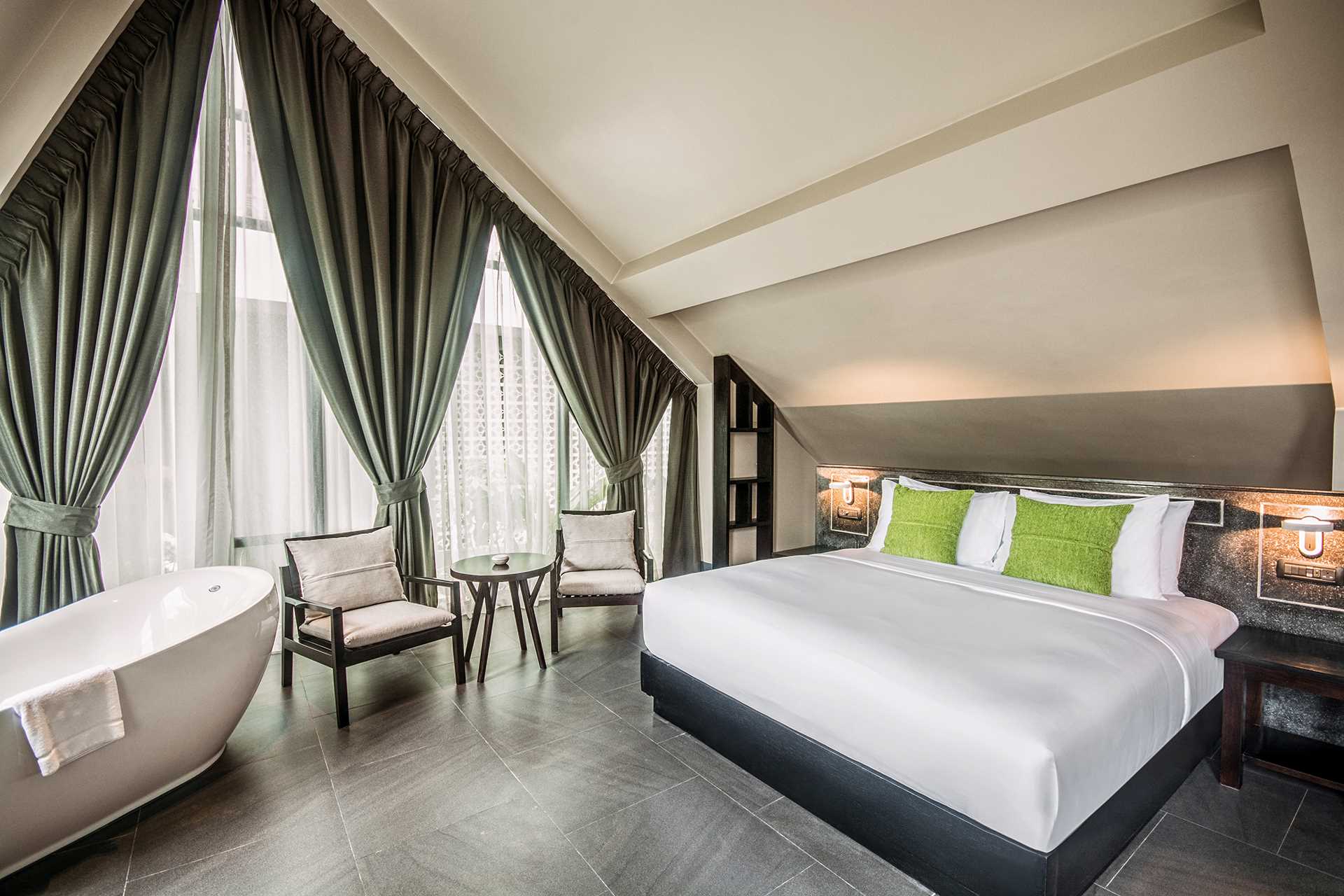 Anjali by Syphon luxurious hotel room in Siem Reap featuring a large bed with green linens, freestanding bathtub near window with curtains, and a seating area with two chairs.